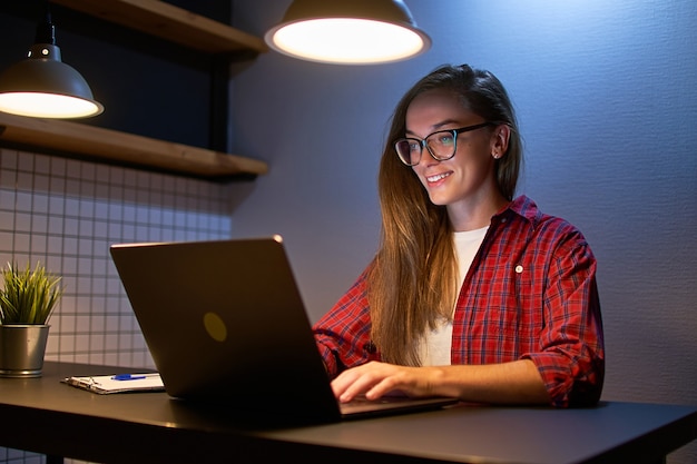 オンラインウェビナーコースを見て、会議のビデオ通話で通信する眼鏡の幸せな若い女の子の学生。夕方に自宅でラップトップを使用して学習するE