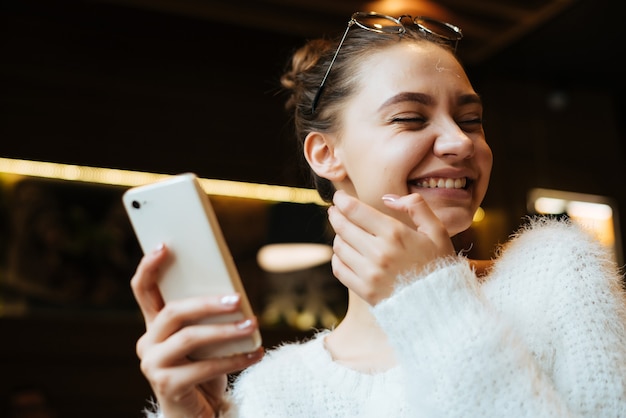 Una studentessa felice con un maglione bianco scrive un messaggio sul suo smartphone e ride