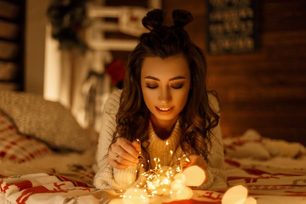 Happy young girl in stylish fashion sweater with festive lights on the bed