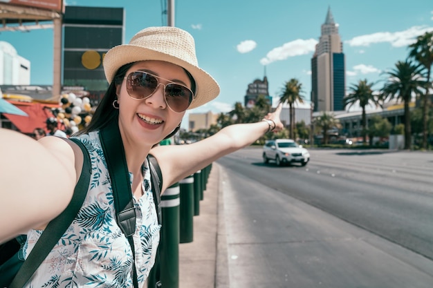 Happy young girl tourist smiling taking self-portrait picture\
on the city center. traveler with hat and sunglasses pointing to\
the tallest building in las vegas on sunny day. travel woman\
lifestyle.