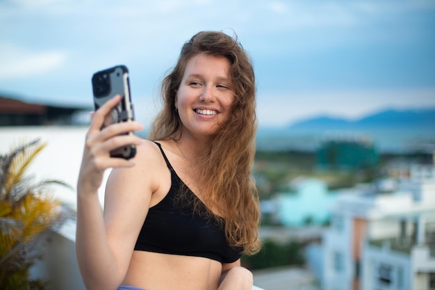 행복한 어린 소녀는 집 발코니 도시 배경의 지붕에서 그녀의 전화기를 사용합니다. 여름에 셀카를 찍는 여성은 휴대폰 스마트폰으로 자신의 재미있는 사진을 가지고 웃으며 화상 채팅을 합니다.