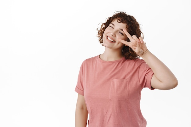 Photo happy young girl with curly hair, winking and smiling with v-sign, positive kawaii gesture near eye, standing upbeat on white