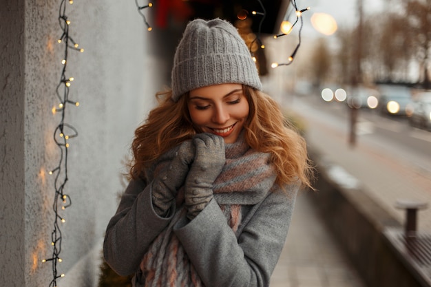 Счастливая молодая девушка с волшебной улыбкой в модной трикотажной одежде в вязаной шапке позирует в городе у фонарей