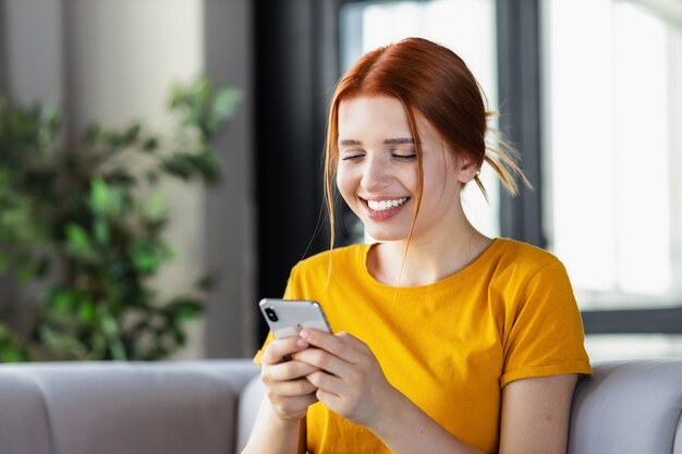 Счастливая молодая девушка с рыжими волосами использует мобильный телефон для онлайн-общения, просмотра забавных видеороликов, социальных сетей, сидя дома на диване, улыбаясь