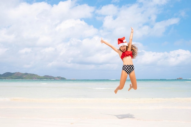 빨간 산타 모자를 쓴 행복한 어린 소녀가 화제의 해변에서 기쁨과 재미를 뛰어넘는 여름 휴가 개념