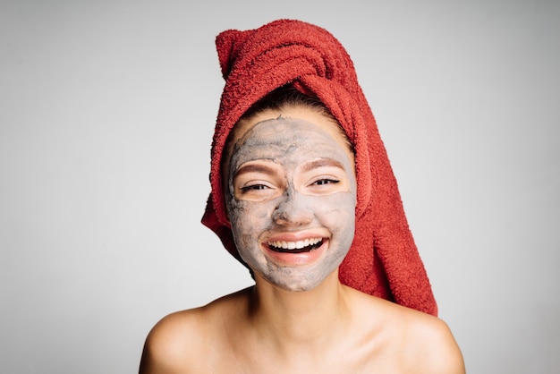 Happy young girl with a red towel on her head pinched a useful clay mask on her face, laughs, a day spa