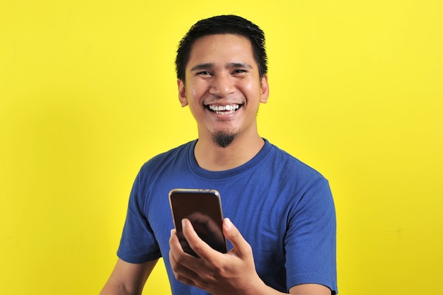 黄色の背景で隔離の携帯電話を使用して笑って幸せな若い格好良いアジア人男性