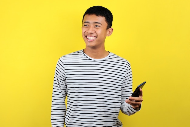 노란색 배경에 격리된 스마트폰을 사용하여 웃고 있는 행복한 젊고 잘 생긴 아시아 남자