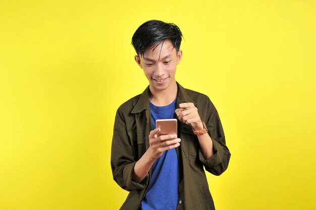 黄色の背景で隔離のスマートフォンを使用して笑っている若い格好良いアジア人男性の幸せ