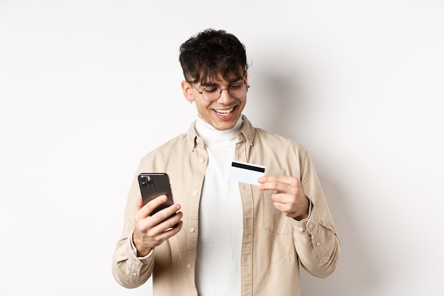 Felice giovane ragazzo in bicchieri che fanno acquisti online, guardando la carta di credito in plastica e tenendo lo smartphone, in piedi sul muro bianco.