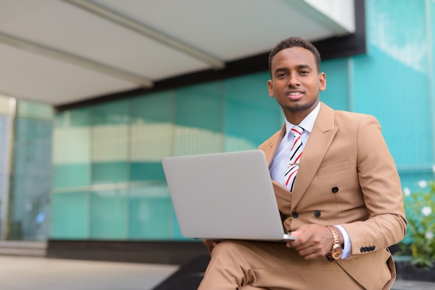 Happy young handsome African businessman with laptop sitting in the city outdoors