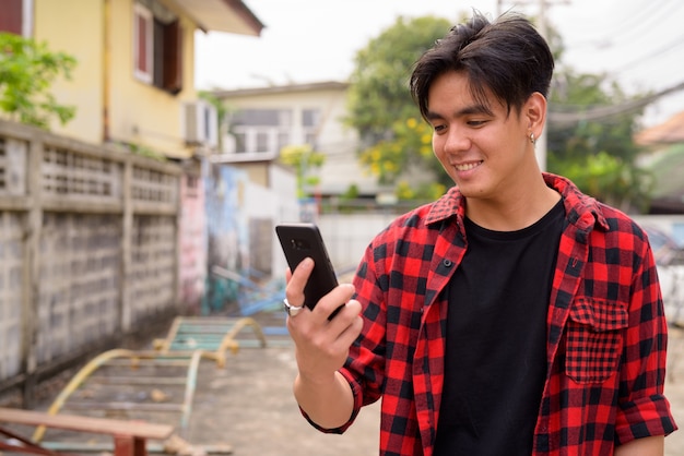 Happy young handsome Asian hipster man using phone in the streets outdoors