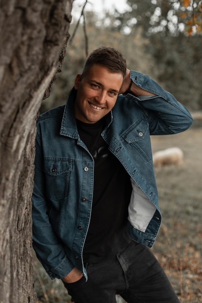 Happy young handsome hipster guy with a smile in a fashionable denim jacket black Tshirt and jeans standing near a tree in the countryside