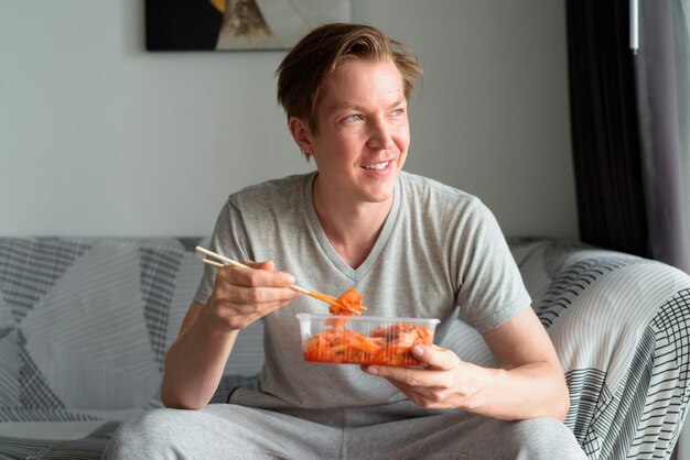 Happy young handsome man eating kimchi and thinking in the living room at home