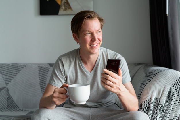 家で考えながらコーヒーカップと電話を保持している幸せな若いハンサムな男