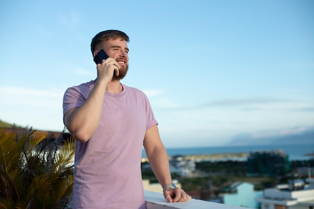 幸せな若いハンサムな男は、夏の晴れた日に屋外で笑顔の友人と会話をしているスマートフォンで呼び出す携帯電話で話しています