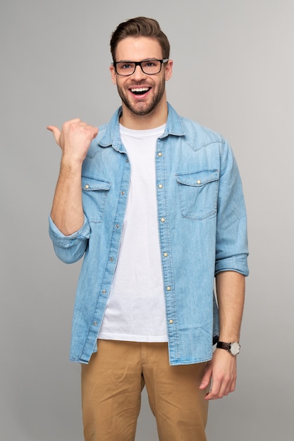 Happy young handsome man in jeans shirt pointing away standing against grey wall