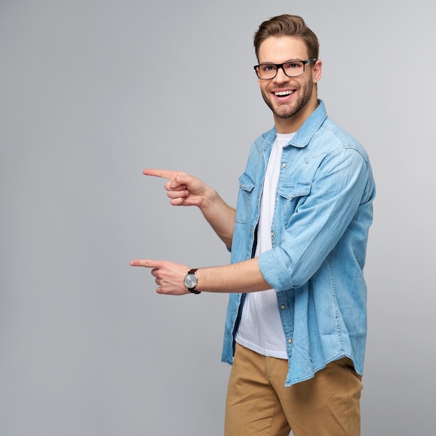 Happy young handsome man in jeans shirt pointing away standing against grey wall