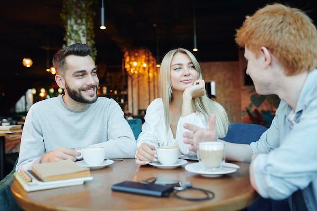 Happy young heterosexual couple chatting to their friend by cup of cappuccino while relaxing in cafe after work or college