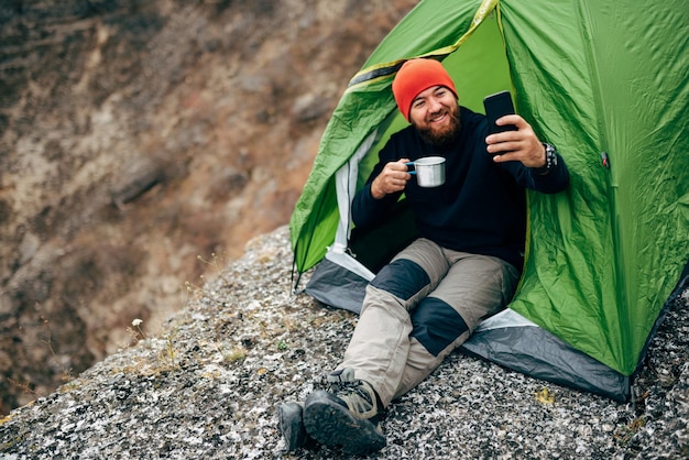 Счастливый молодой турист мужчина улыбается, делая селфи в горах со своего смартфона Путешественник бородатый мужчина в красной шляпе делает автопортрет с мобильного телефона после похода