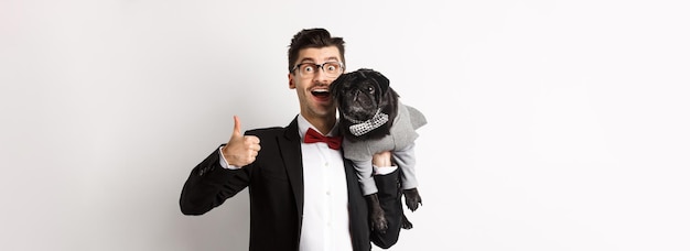 Happy young hipster in suit and glasses showing thumbup holding cute black dog on shoulder love his