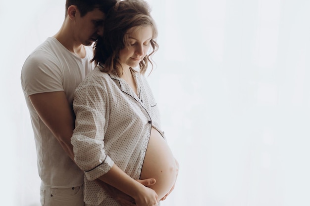 Happy young husband hugging his pregnant wife and holding belly bump Stylish pregnant family mom and dad in white pajamas relaxing at home in morning light Fertility concept True happiness