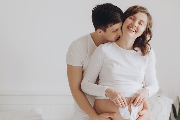 Happy young husband kissing his pregnant smiling wife and holding white little socks on belly bump Stylish pregnant family mom and dad in white relaxing at home and hugging belly f