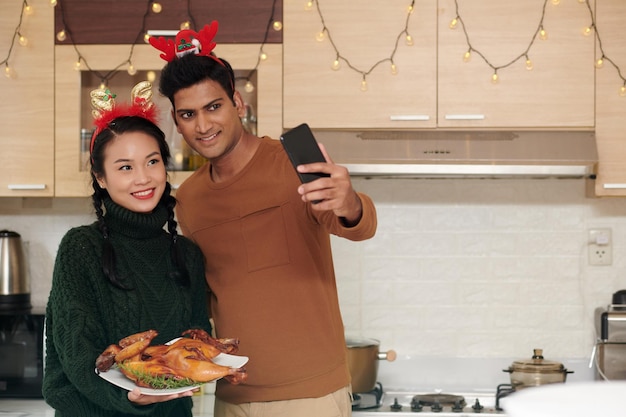 Happy young husband and wife with reindeer antlers headband taking selfie and showing grilled chicke...