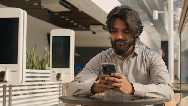 Happy young indian businessman holding smartphone sending message sitting indoors smiling arabian