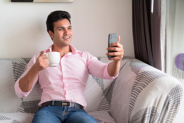 Счастливый молодой индийский бизнесмен, делающий селфи, пить кофе дома