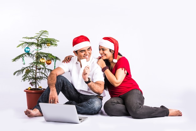 Happy and young Indian couple on sofa using laptop while celebrating Christmas