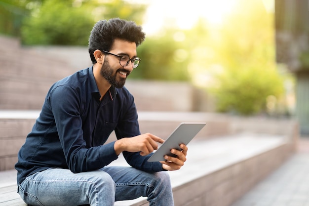 Happy young indian guy sitting on stairs using digital tablet