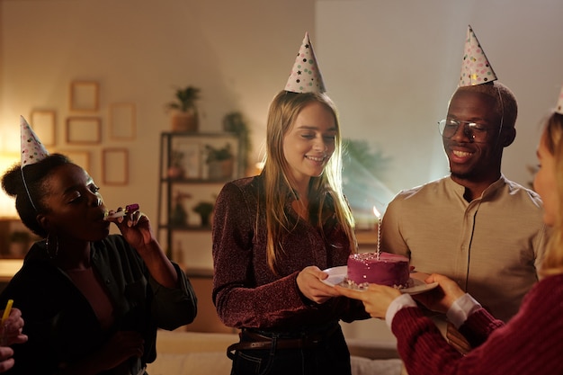 happy young intercultural friends congratulating gorgeous blond girl on her birthday while pretty female passing her plate with delicious cake