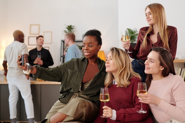 Happy young intercultural women with flutes of champagne making selfie while cheering up and enjoying home party against their boyfriends