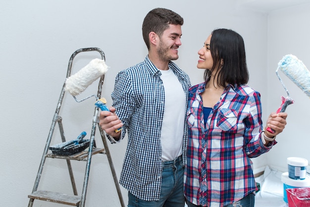 Happy young interracial couple looking each other and holding\
roller to paint themselves his new apartment.