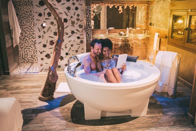 Happy young interracial couple relaxed enjoying their self in the jacuzzi drinking wine and with a tablet