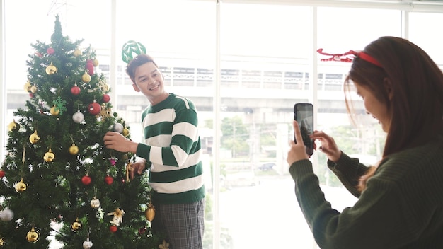 Happy young joyful Asian couple having fun taking photos decorating baubles on the Christmas tree