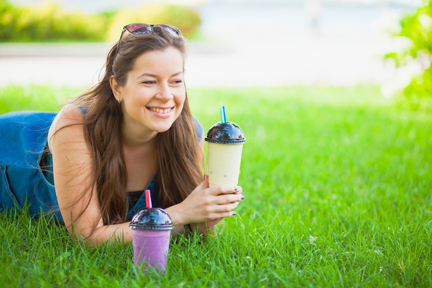 草の上の幸せな若い女性は、晴れた夏の日にジューシーなスムージーを飲みます。