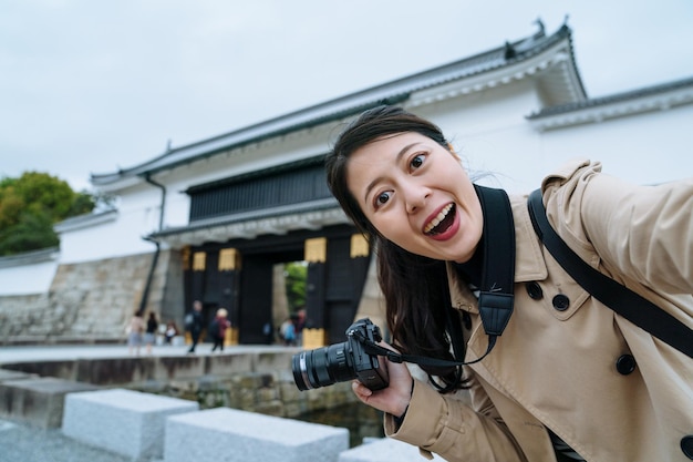 カメラを持った幸せな若い女性が、二条城の入り口でスマートフォンでセルフィーを撮っています。カメラに面したアジアの女性旅行者は、日本の有名な歴史的建造物を見て興奮しています。