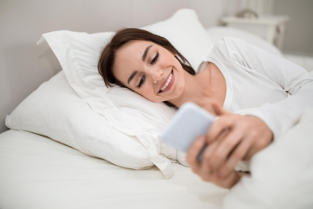 Happy young lady holding smartphone while lying in bed