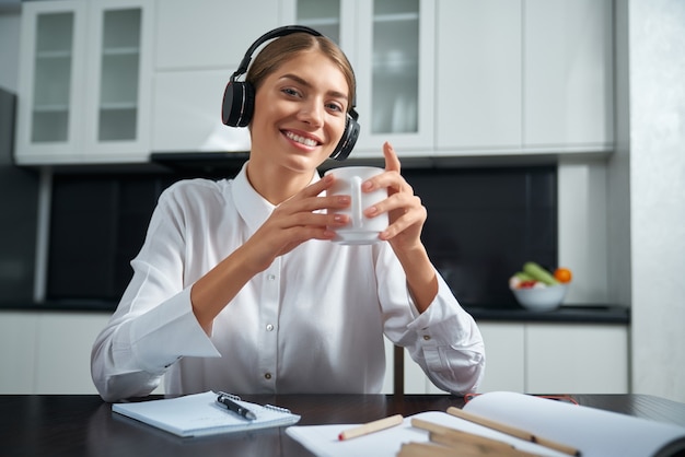 Happy young lady in wireless headphones sitting at kitchen table and drinking hot tea