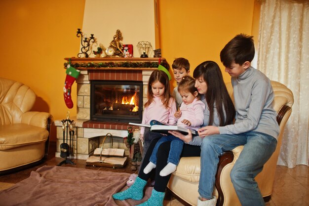 Happy young large family by a fireplace in warm living room on winter day Mother with four kids at home read book