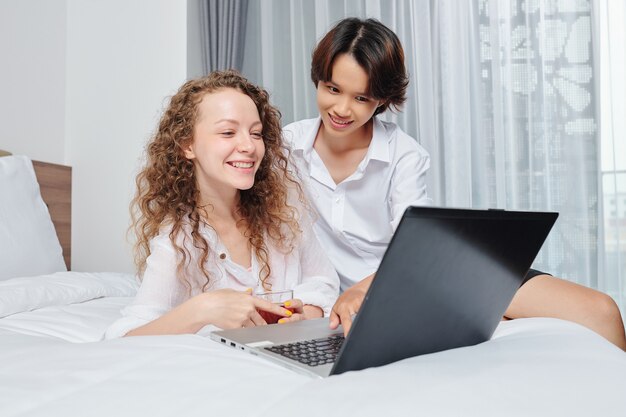 Happy young lesbian couple resting in bed with cups of tea and watching movie on laptop screen