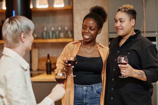 Happy young lesbian couple with glasses of red wine chatting to their friend while standing in front