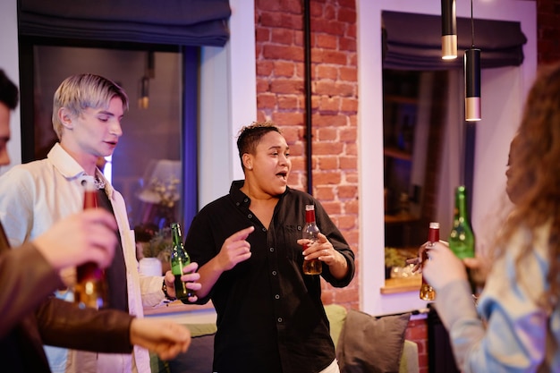 Happy young lgbtq people with bottles of beer dancing in living room in the evening while enjoying h