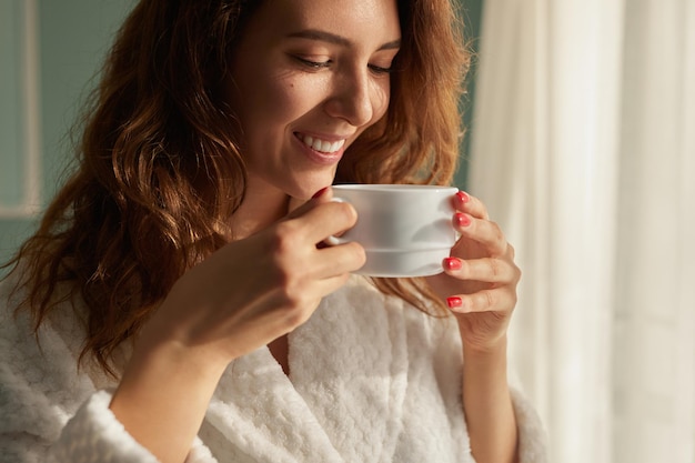 Happy young long haired female in homey robe enjoying fresh aromatic coffee from white mug while standing near window in sunny morning at home