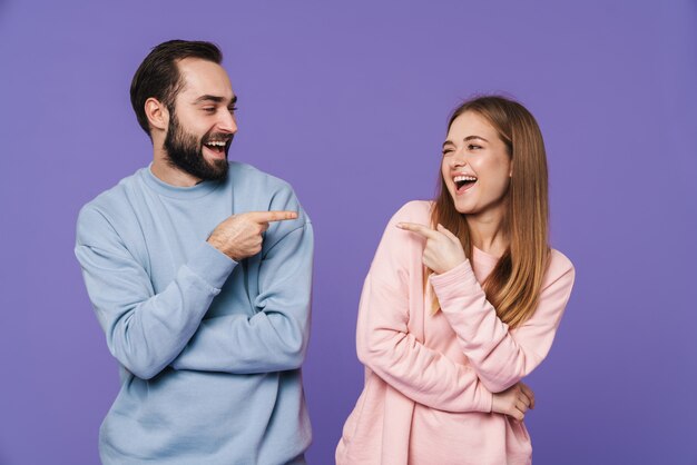 a happy young loving couple isolated over purple wall pointing to each other.