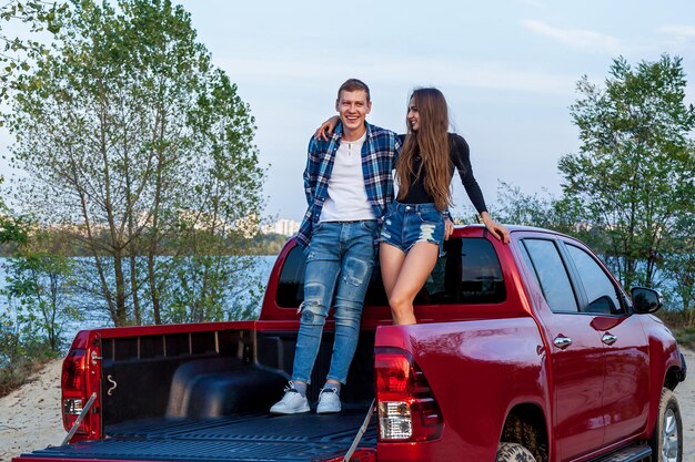 湖の近くのビーチで赤い車の後ろに笑みを浮かべて抱き締めて幸せな若い愛情のあるカップル