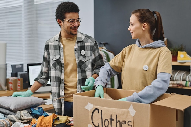 Happy young male and female volunteers chatting while packing box with clothes