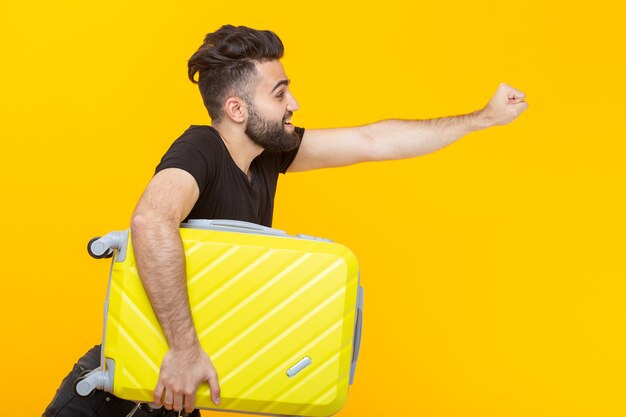 Happy young male hipster with a beard holding a yellow suitcase on a yellow background and rejoicing